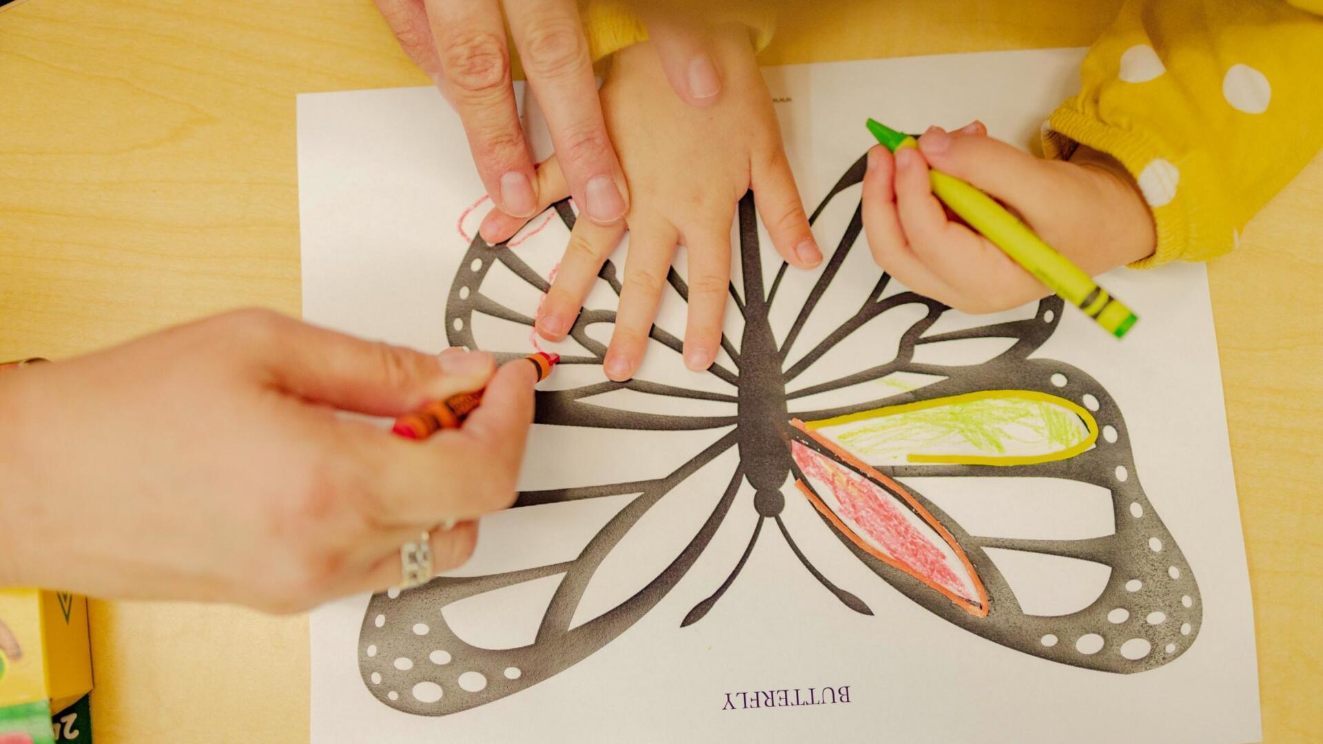 An adult’s hands guide a child’s hands while coloring a butterfly outline on a sheet of paper with crayons.